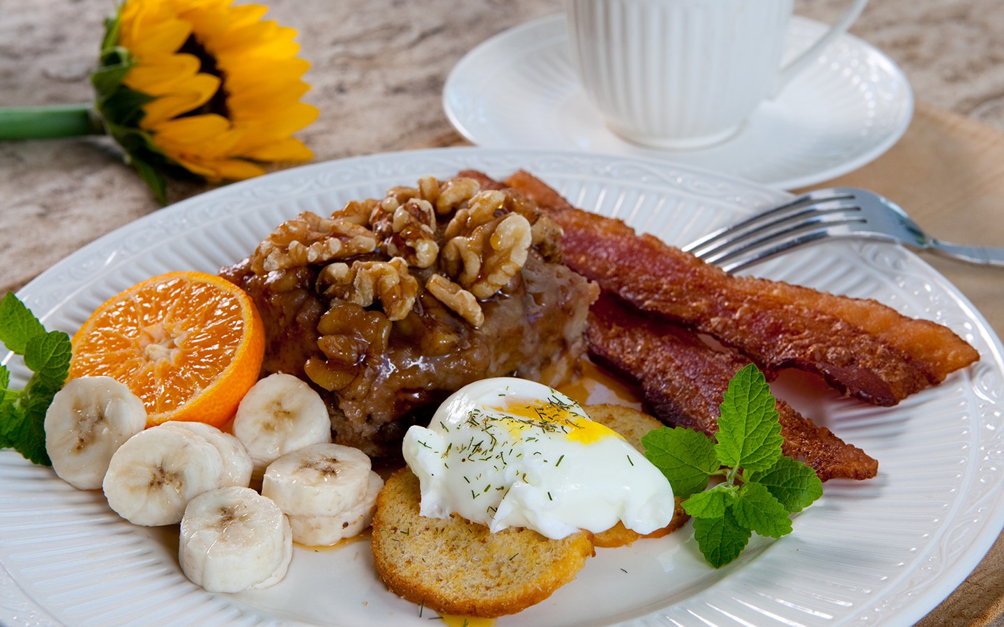 White plate with bacon, poached egg on toast, sliced banana, orange half with pastry covered with walnuts and coffee mug.