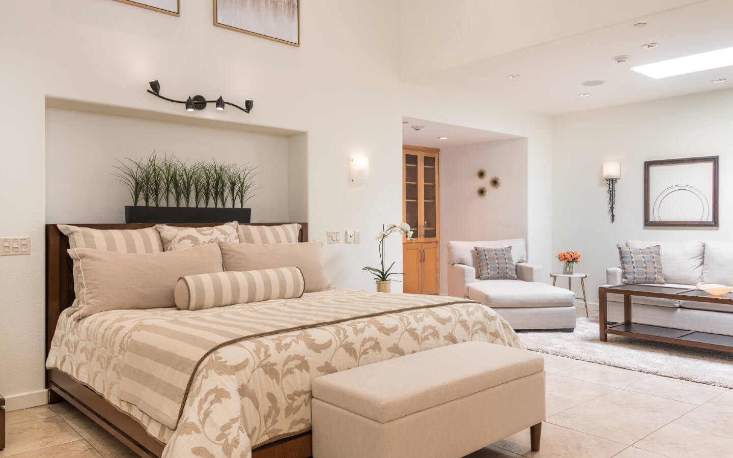 Bedroom with recessed bed with beige and tan pillows. Bamboo grows in recess above bed, underneath artwork. Cabinet and stuffed chair sit in alcove.