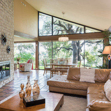 Sunny living room with tall sloped ceiling. Leather L-shaped sofa and coffee table accent sitting area across from brick fireplace.