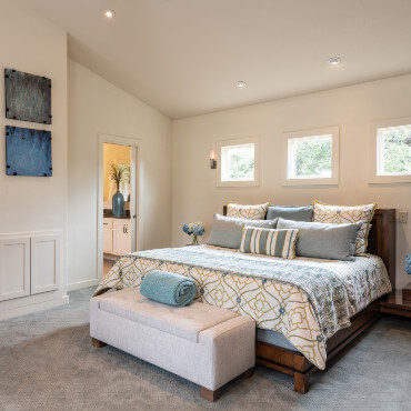 High walled sloped bedroom with three windows above a bed. Open door to bathroom with decorative vase, next to modern artwork on wall.