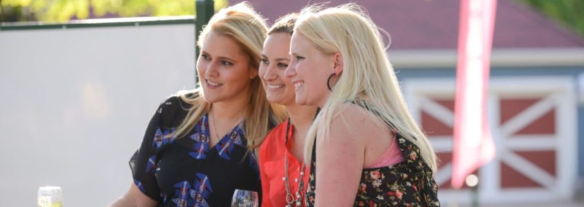 Three women sitting on a bench outside smiling and holding wine glasses