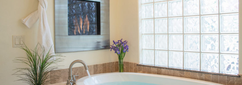 Framed fireplace recessed into wall above jetted tub filled with water, underneath cloudy opaque glass block window. Flowers decorate the tub area.