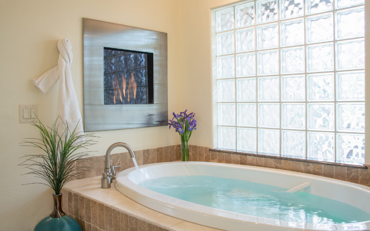 Framed fireplace recessed into wall above jetted tub filled with water, underneath cloudy opaque glass block window. Flowers decorate the tub area.