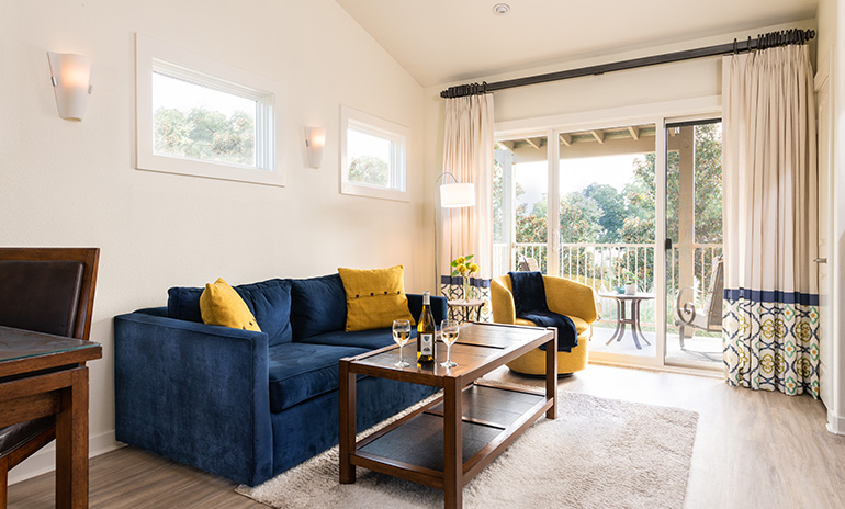 View over living area with blue couch and sunflower yellow accents, and yellow barrel chair with blue accents, and out the open wall to wall patio doors to seating area outside with view of trees in background.