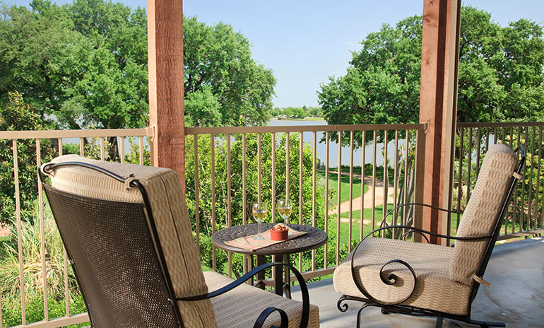 Outdoor wicker padded seating area with decorative patio table holding wine glasses and snack as patio offers view of waterfront and trees.