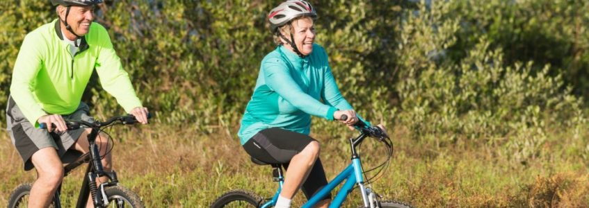 Older man and woman wearing helmets and biking a trail surrounded by natural landscaping